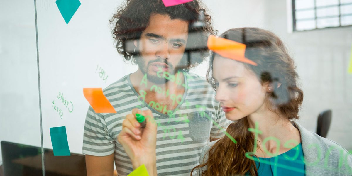 Estrategia-inbound-marketing-destaque Homem e mulher escrevem em papeis coloridos colados a um vidro. Estão desenhando uma estratégia inbound marketing.