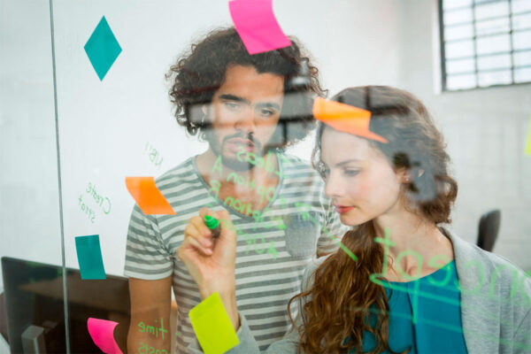 Estrategia-inbound-marketing-destaque Homem e mulher escrevem em papeis coloridos colados a um vidro. Estão desenhando uma estratégia inbound marketing.